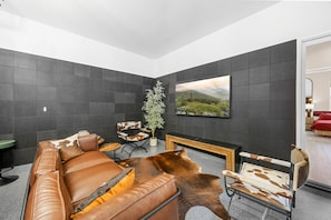 A modern living room with a leather sofa, a coffee table, a fireplace, and a large landscape painting on the wall. - Design Estate w/Heated Zero-Edge Pool & Game Room (Coachella)