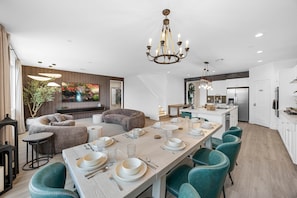 A modern kitchen with white cabinets, a central island, and a dining area with a long table set for six. - Design Estate w/Heated Zero-Edge Pool & Game Room (Coachella)