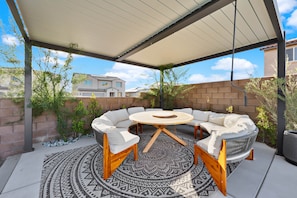 A covered patio area with a round wooden table and white cushioned seating. - Design Estate w/Heated Zero-Edge Pool & Game Room (Coachella)