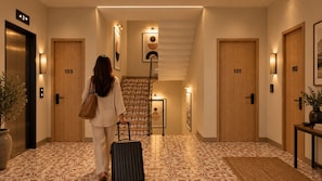 Un pasillo de hotel con una persona arrastrando una maleta, puertas con los números 101 y 103, y una escalera que lleva a un nivel superior.
