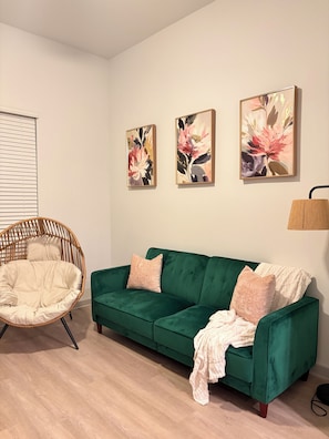 Ein Zimmer mit einem grünen Sofa, einem Korbstuhl und zwei floralen Bildern an der Wand.