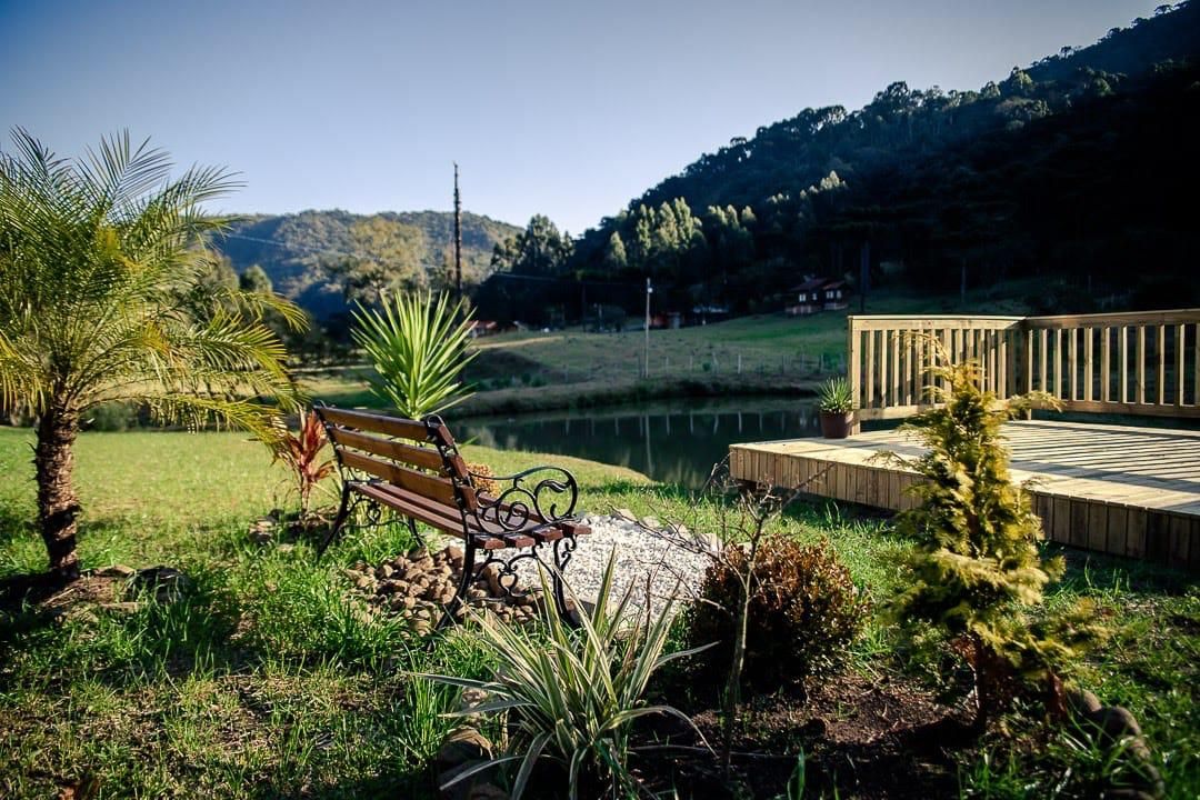 Um deck de madeira com um banco, rodeado por vegetação e vista para um lago e colinas.