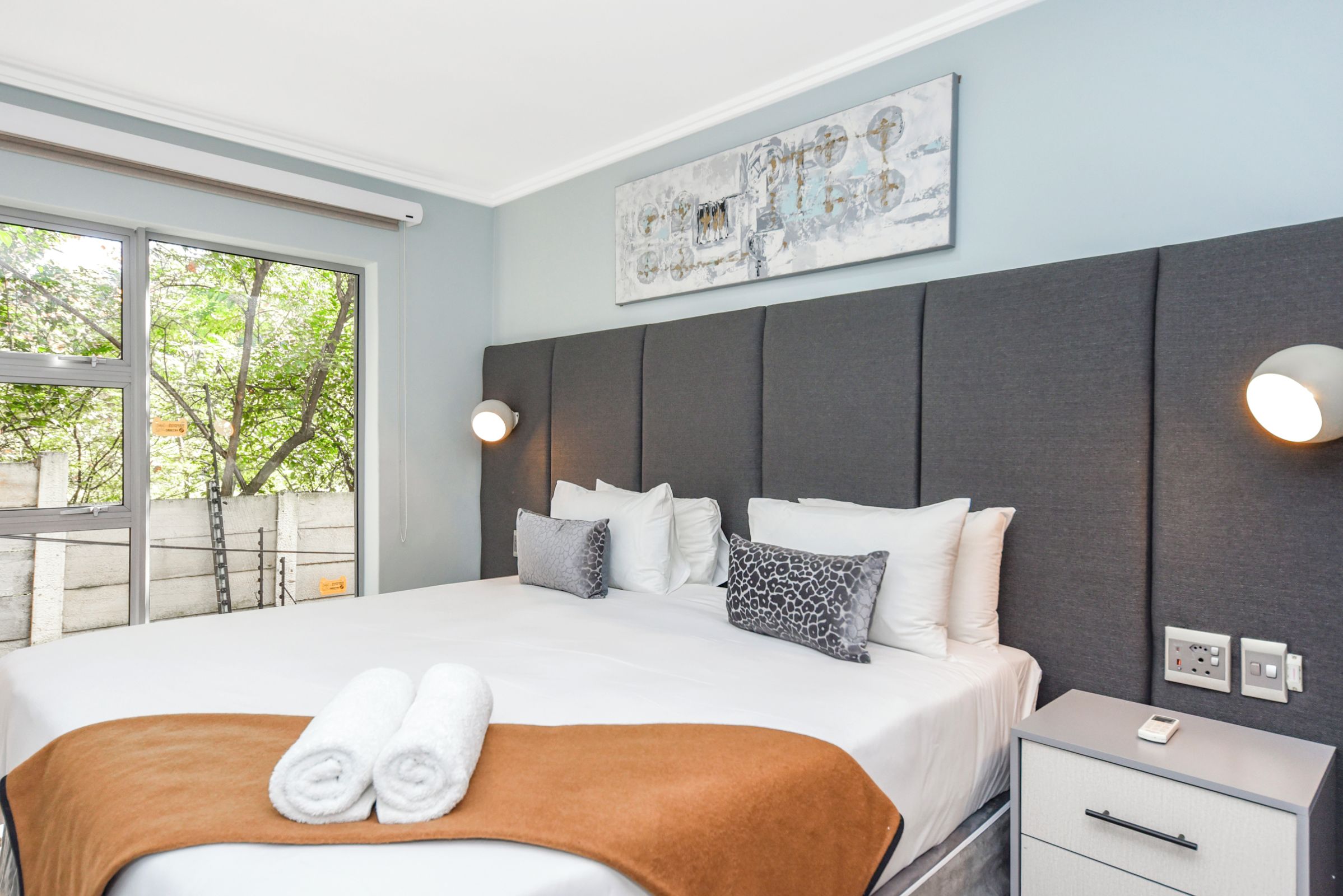 Une chambre d’hôtel avec un grand lit, une tête de lit, une table de chevet, une lampe fixée au mur et une fenêtre donnant sur des arbres.