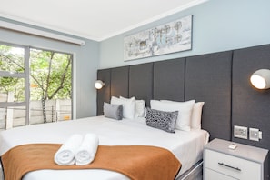 A hotel room with a large bed, a headboard, a nightstand, a wall-mounted light, and a window with a view of trees. - Luxury 2Bed Retreat Sandton With Free Parking (Sandton)