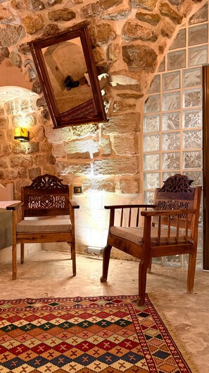 A room with stone walls, two wooden chairs with intricate carvings, a patterned rug, and a large mirror on the wall. - YONA OTEL (ARTUKLU)