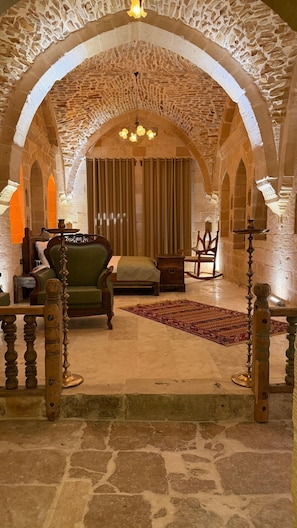 A room with a stone arch ceiling, a green sofa, a wooden chair, a small table, and a patterned rug. - YONA OTEL (ARTUKLU)