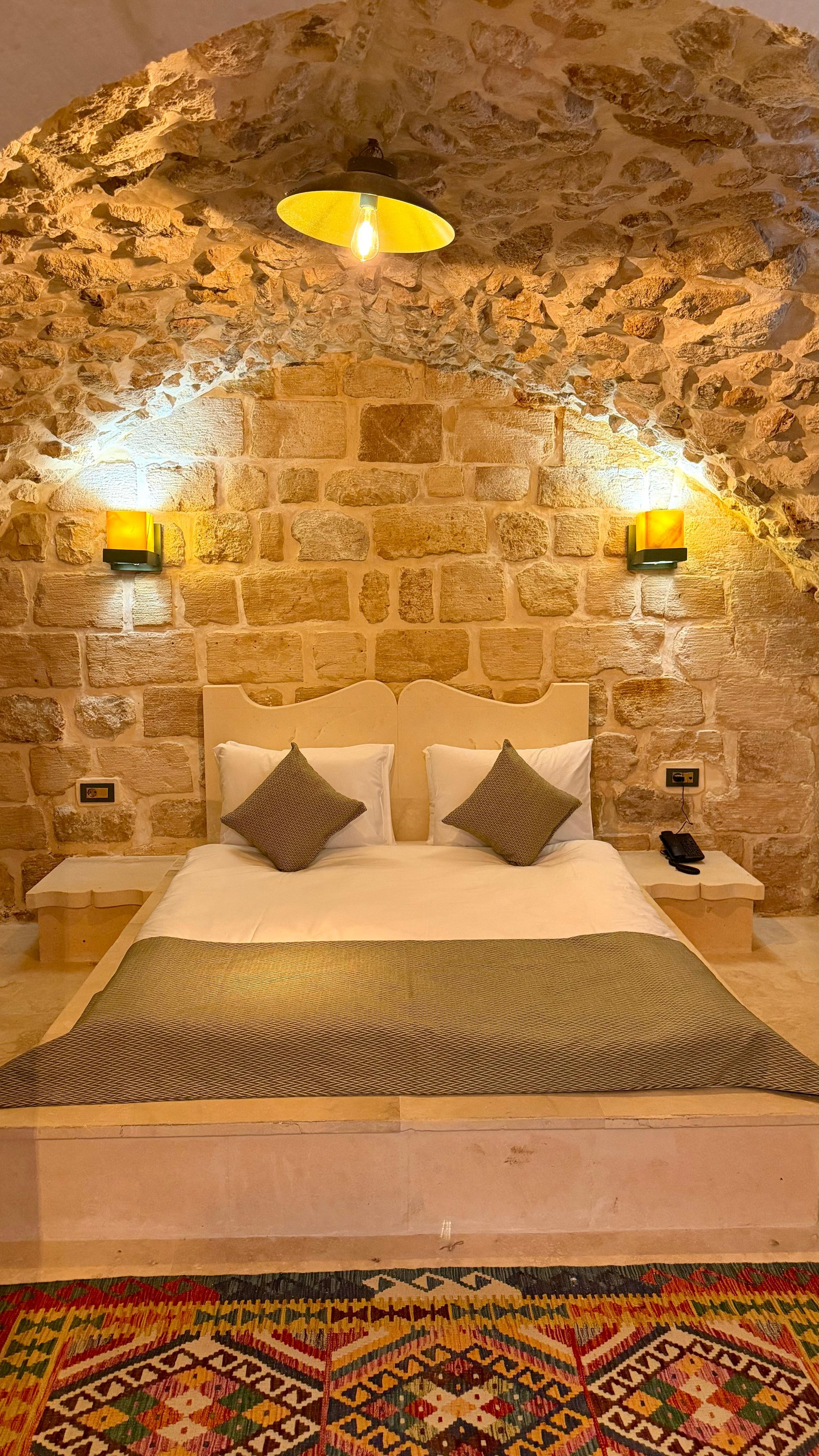 Una habitación con pared de piedra, cama con ropa de cama blanca, dos lámparas de noche y una alfombra estampada.