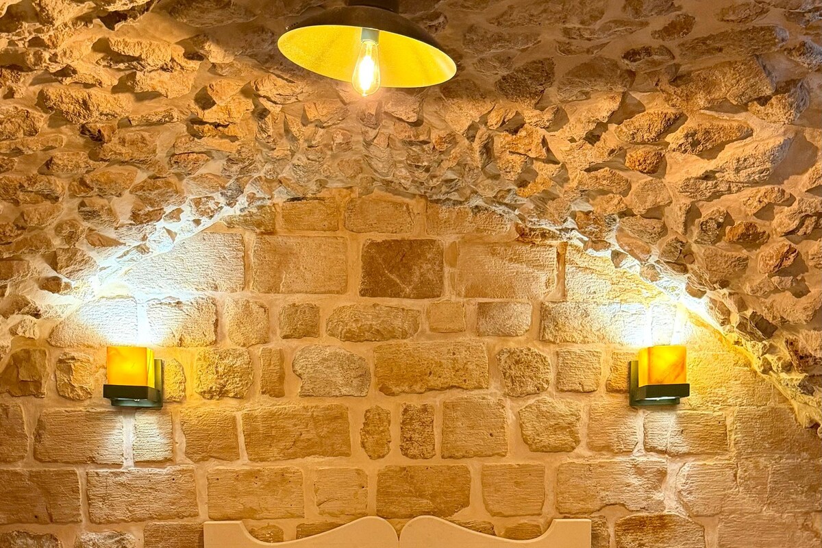 Una habitación con pared de piedra, cama con ropa de cama blanca, dos lámparas de noche y una alfombra estampada.