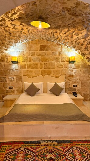 Una habitación con pared de piedra, cama con ropa de cama blanca, dos lámparas de noche y una alfombra estampada.
