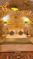 Una habitación con pared de piedra, cama con ropa de cama blanca, dos lámparas de noche y una alfombra estampada.