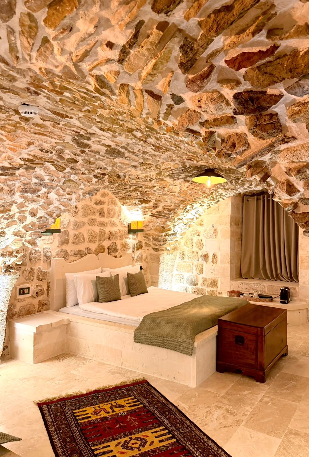 Un dormitorio con paredes de piedra, una cama, una lámpara de noche y un cofre de madera.