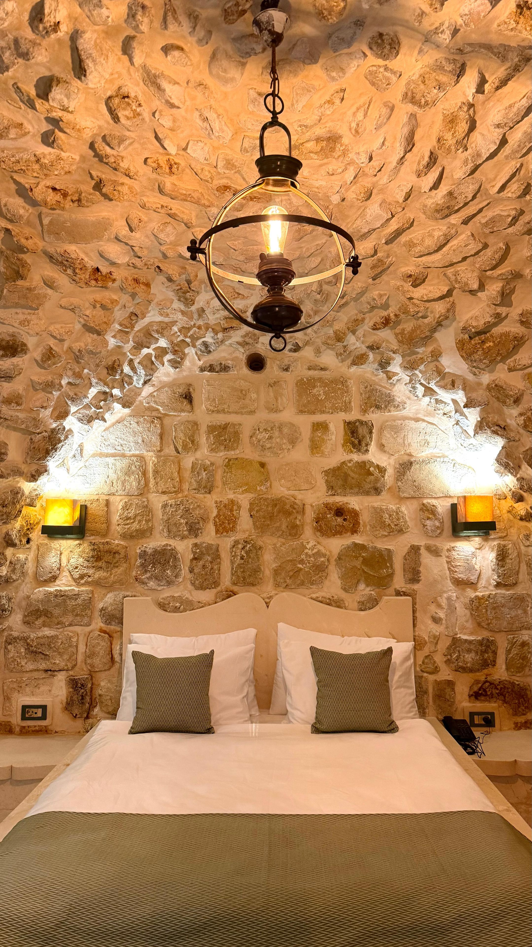 Una habitación con pared de piedra, una cama con dos almohadas y una lámpara colgante.