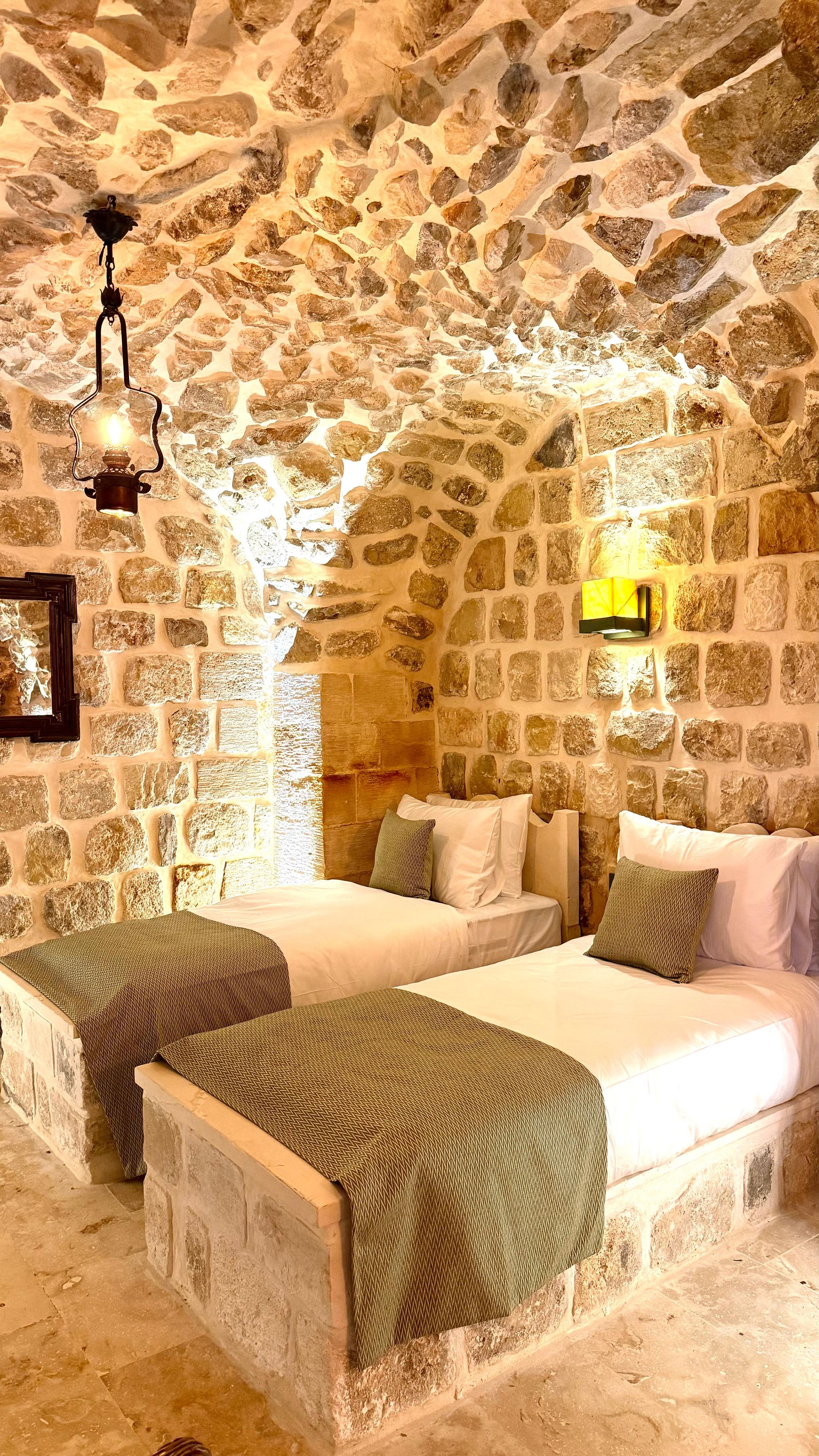 Habitación de paredes de piedra con dos camas, una lámpara colgante y una lámpara de pared.