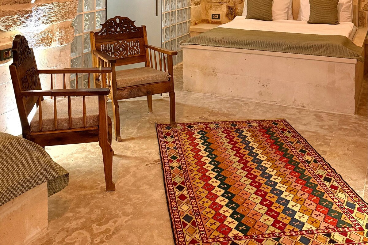 Una habitación con techo de piedra, cama con ropa blanca, una silla de madera y una alfombra colorida.