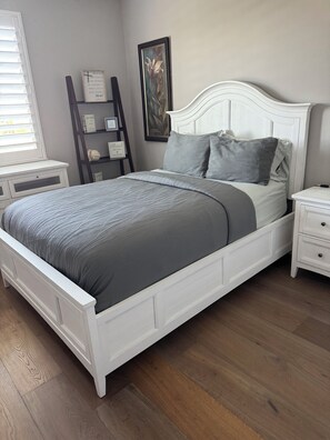 A bedroom with a white bed, grey bedding, a wooden floor, a nightstand, a dresser, a window with blinds, and a wall with a framed picture and a shelf with items. - Cozy 1-bedroom apartment in perfect Winchester with AC (Winchester)
