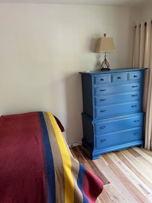 Ein Zimmer mit einem Bett, das mit einer Streifenauflage bedeckt ist, einer blauen Kommode mit Schubladen, einer Lampe darauf und einem Fenster mit Vorhängen.