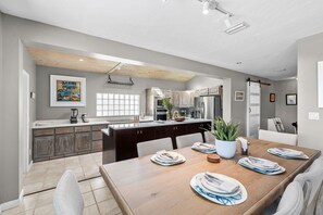 A modern kitchen with a central island, a dining area with a wooden table set for four, and a large window providing natural light. - 6 Country Club Rd (Cocoa Beach)