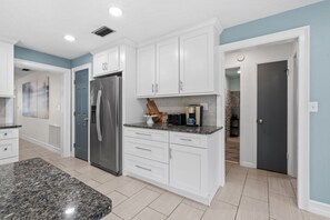 A modern kitchen with white cabinetry, a stainless steel refrigerator, a granite countertop, and a coffee maker. - 122 Sunset Dr (Cocoa Beach)