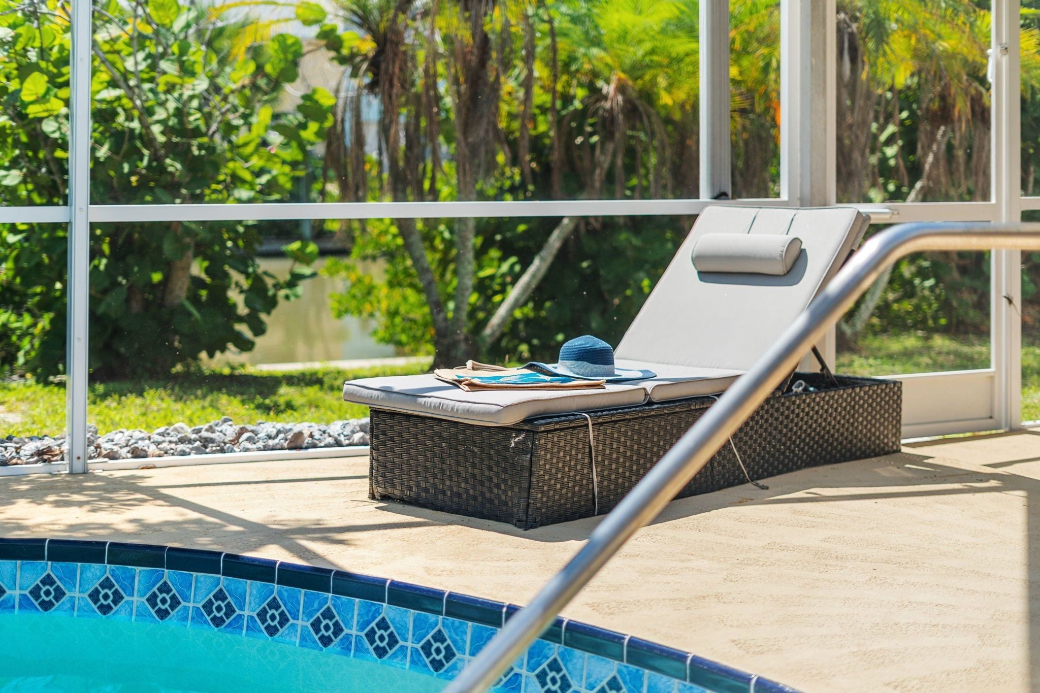Una silla de descanso junto a la piscina con cojín, un sombrero, una cesta de mimbre y un borde de piscina con azulejos.