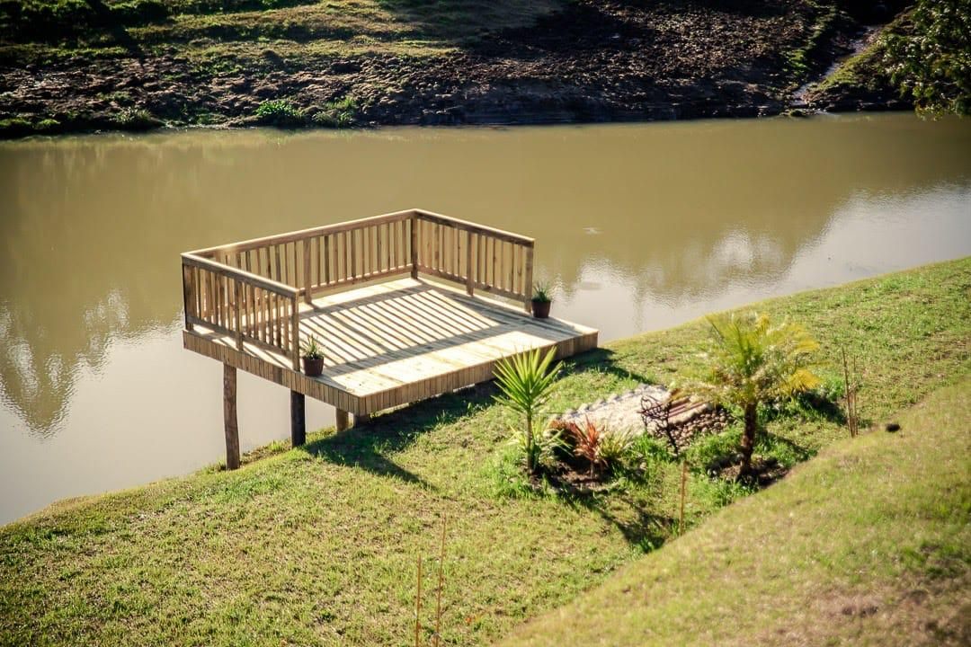 Una terraza de madera que se extiende sobre un cuerpo de agua tranquilo, rodeada de orillas cubiertas de hierba y plantas pequeñas.