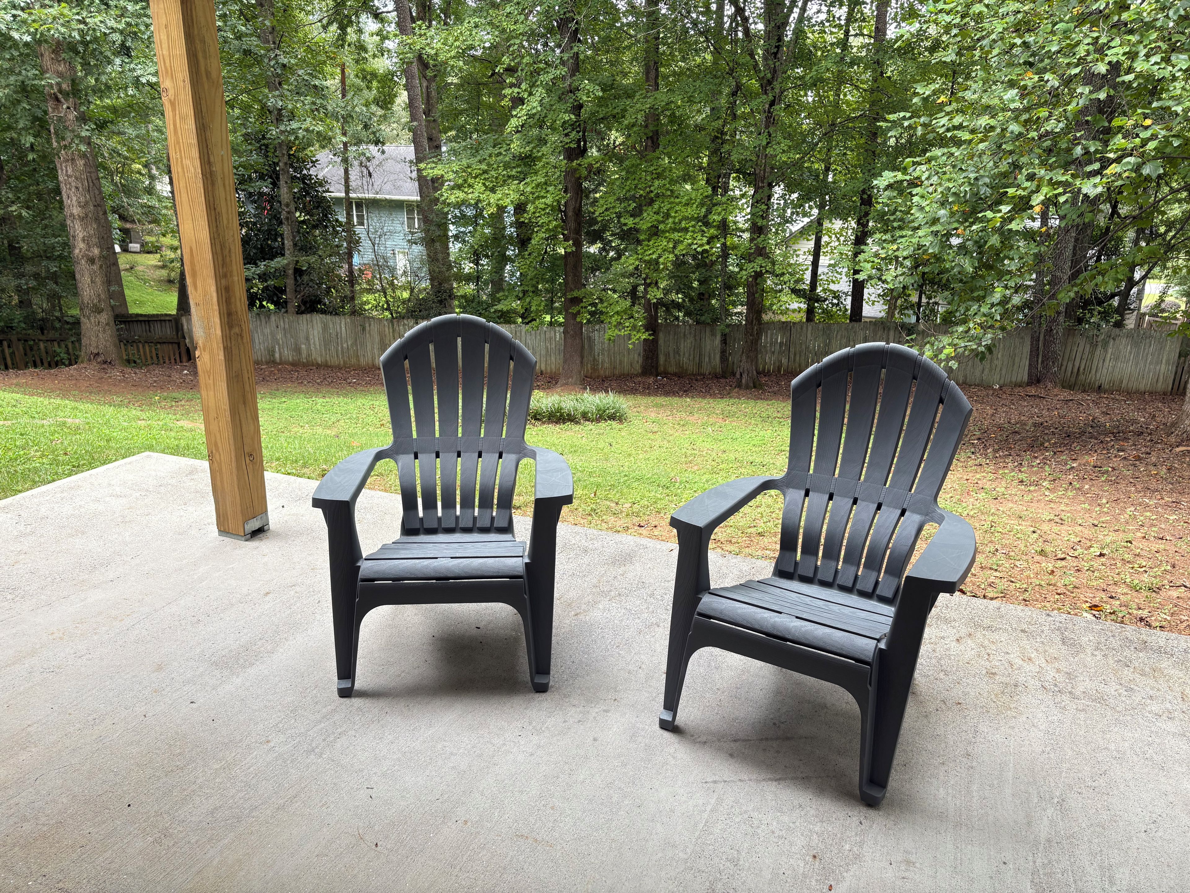 Two Adirondack chairs on a concrete patio with a wooden post nearby, surrounded by a grassy yard and a wooden fence.