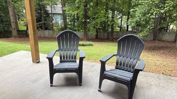 Two Adirondack chairs on a concrete patio with a wooden post nearby, surrounded by a grassy yard and a wooden fence.