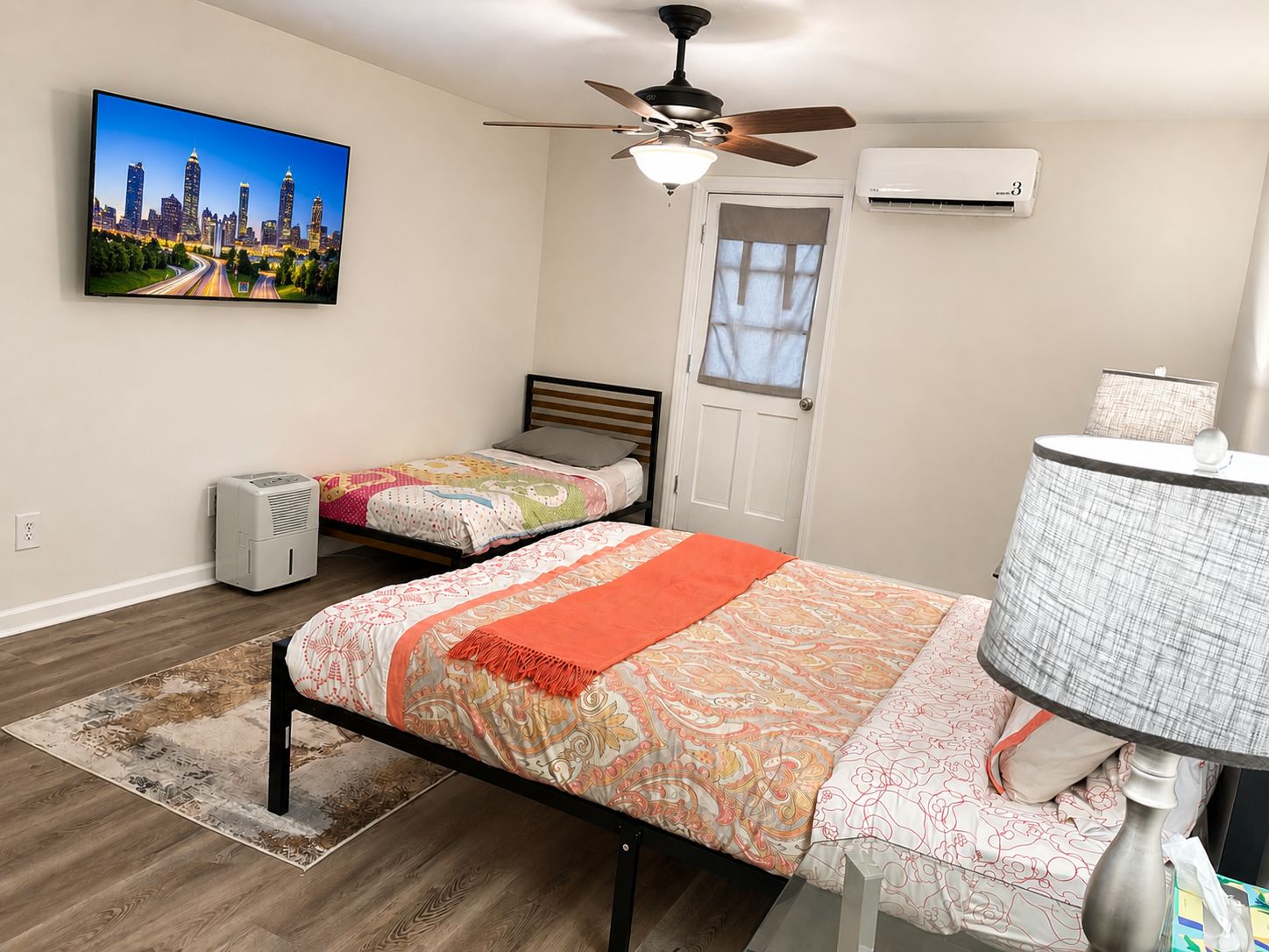 A bedroom with two beds, a ceiling fan, an air conditioning unit, a desk with a lamp, and a cityscape painting on the wall.