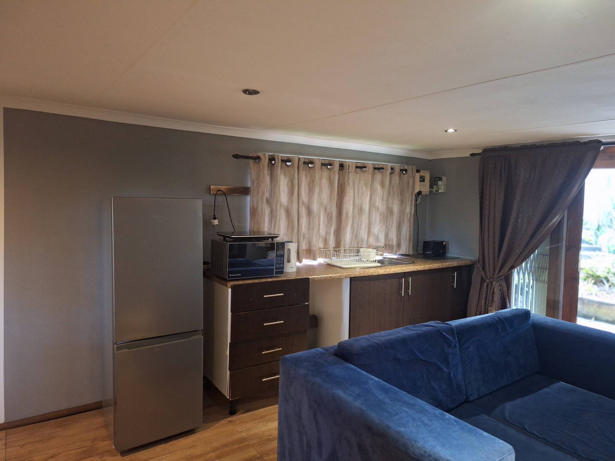 A compact kitchen with a refrigerator, microwave, and a small sink. There is a blue sofa in the foreground and a window with curtains.