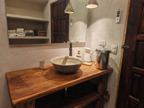 Ein Badezimmer mit einem Holzbadezimmerhocker, einem großen Waschbecken, einem Handtuchspender, einer Kaffeemaschine und einer wandmontierten Leuchte.