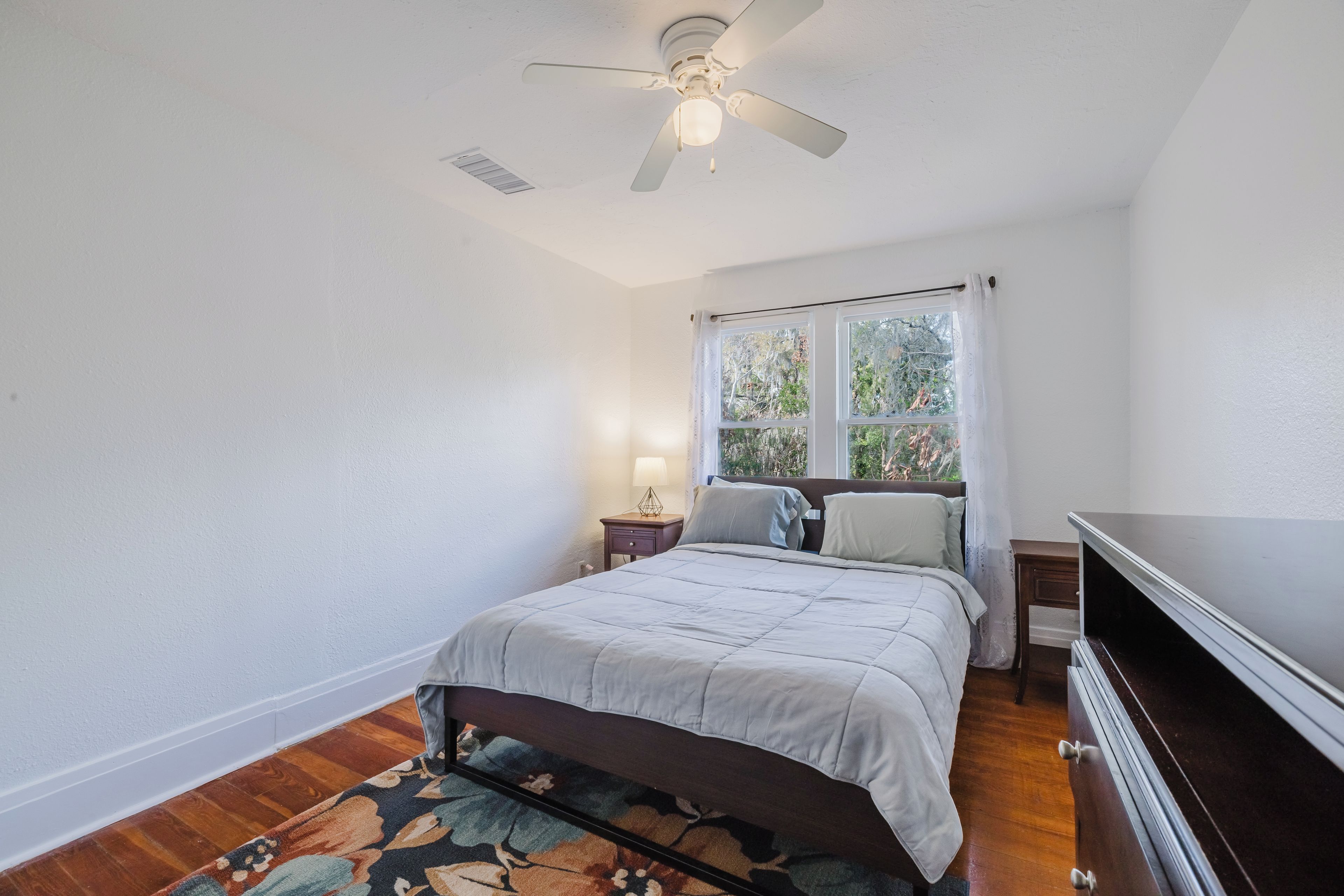 Une chambre à coucher comprenant un lit, une table de chevet, une lampe, un ventilateur de plafond et une fenêtre avec des rideaux.