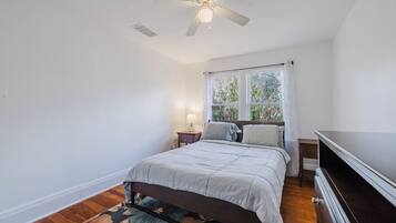 Quarto com cama, mesa de cabeceira, luminária, ventilador de teto e janela com cortinas.