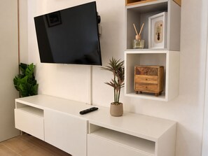 A modern living room with a mounted TV, a white entertainment unit, a potted plant, and a small wooden cabinet. - New Listing (Roma)