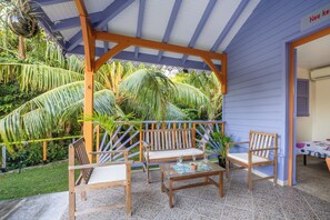 A covered outdoor seating area with wooden furniture and a glass table, surrounded by lush tropical plants. - New Listing (Saint-Pierre)