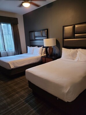 Ein Hotelzimmer mit zwei Betten, einem Nachttisch mit Lampe, einem Deckenventilator und einem Fenster mit Vorhängen.