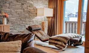 A room with a stone wall, a wooden floor, a lounge chair, a bench, and a window with a view of buildings. - PREMIUM ALPINE RESIDENCE WITH DESIGNER KITCHEN AND HOTEL SPA ACCESS (Zermatt)