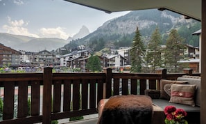 A balcony with a view of a mountain town, wooden railings, and a sofa with cushions. - PREMIUM ALPINE RESIDENCE WITH DESIGNER KITCHEN AND HOTEL SPA ACCESS (Zermatt)