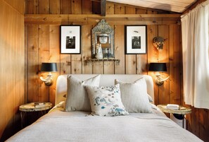 A bedroom with a wooden headboard, a bed with striped bedding, decorative pillows, wall-mounted lamps, and framed pictures on the wall. - Whidbey Island Water View Oasis with Hot Tub. (Freeland)