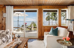 A cozy room with a wooden interior, a white sofa, a small table with a vase of flowers, and a view of the sea through large windows. - Whidbey Island Water View Oasis with Hot Tub. (Freeland)