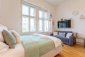 A bedroom with a bed, a grey sofa, a TV, and a window with a view of buildings. - Nově zrekonstruovaný byt vedle Národního muzea (Praha 1)