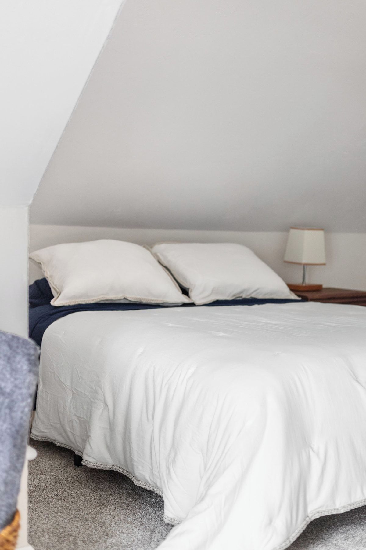 Un dormitorio con cama, almohadas, una mesita de noche y una lámpara.