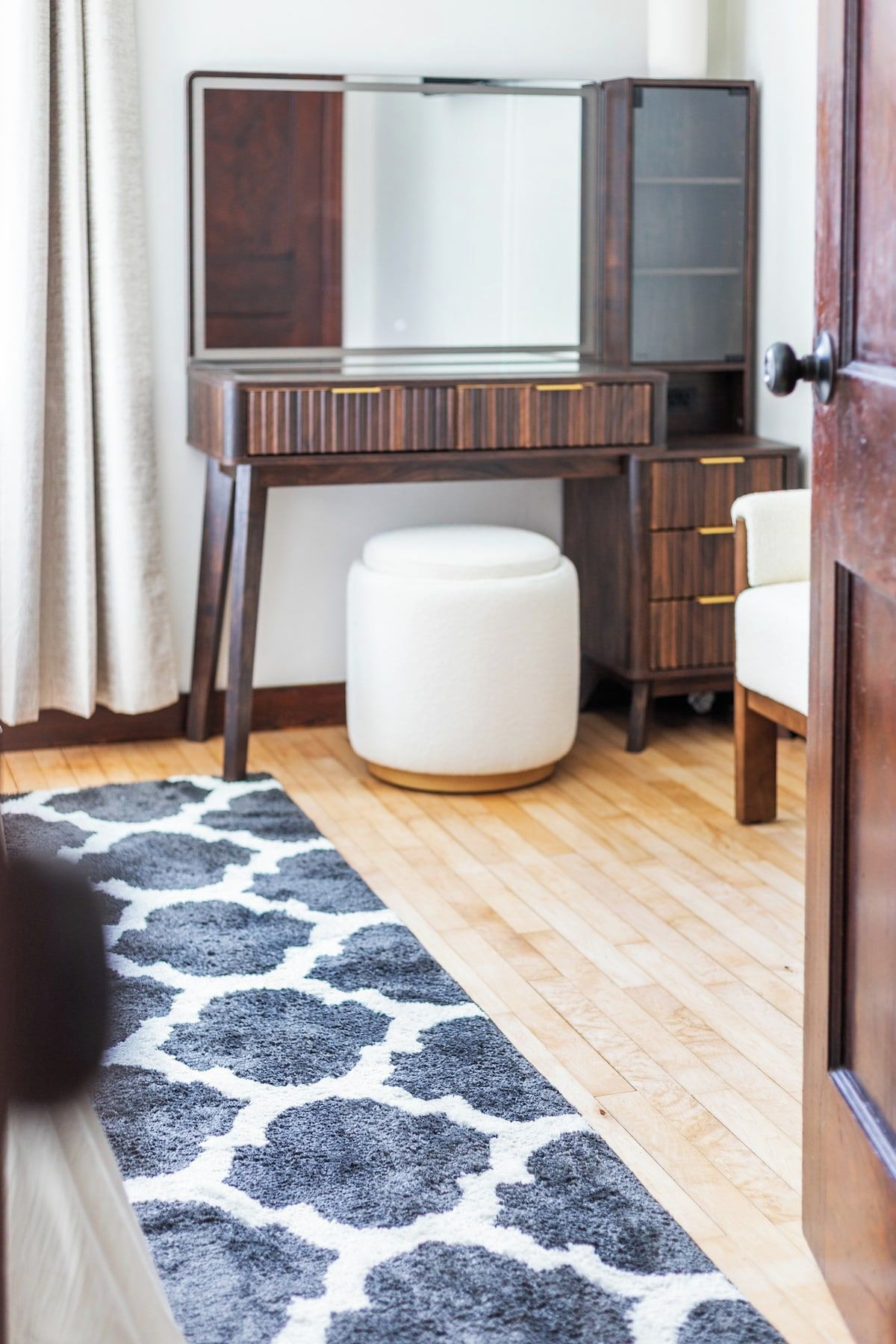 Una habitación con un escritorio de madera, un reposapiés blanco y una alfombra estampada.