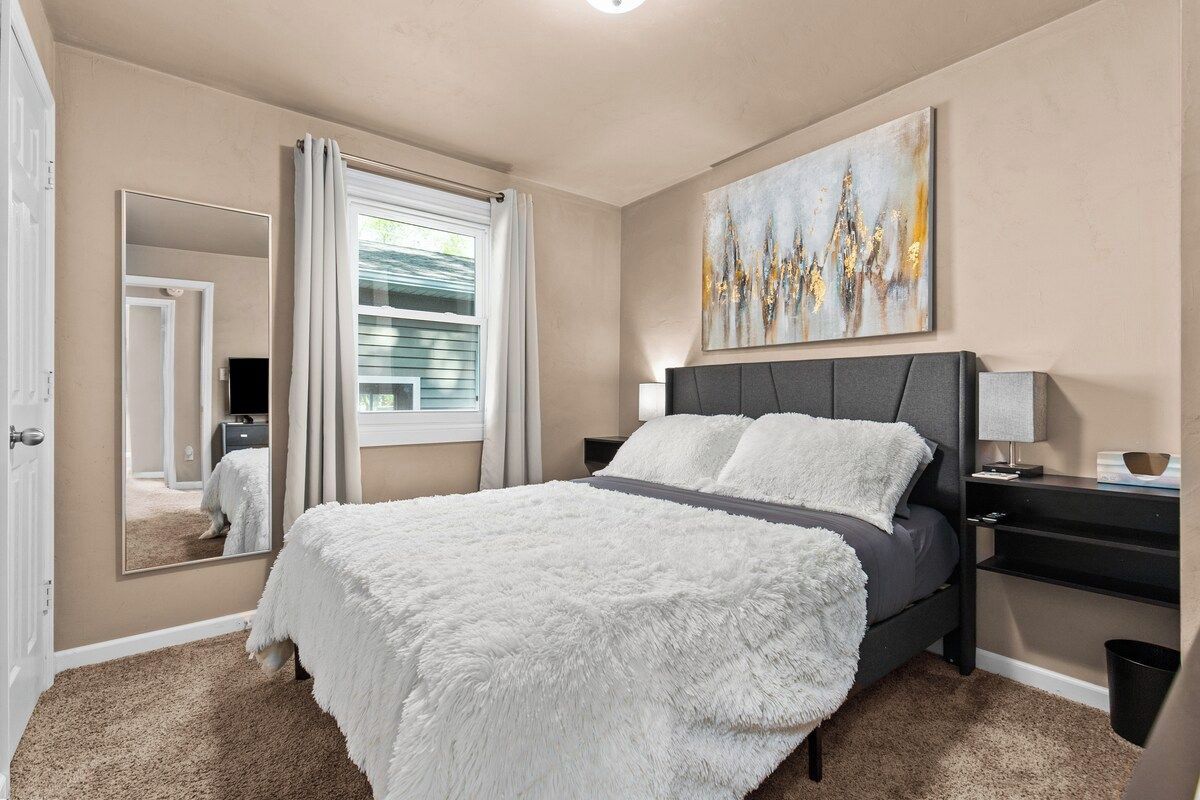 A bedroom with a bed, a nightstand, a mirror, a window with curtains, and a painting on the wall.