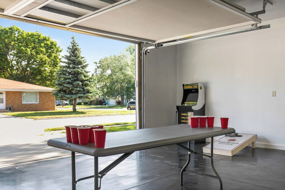 A table with red cups and a dartboard in a garage with a view of a tree and a building outside.