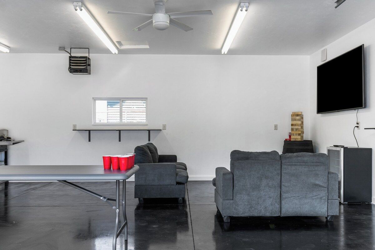 Una habitación moderna con televisión de pantalla plana, ventilador de techo, ventana con persianas y una mesa con vasos rojos.