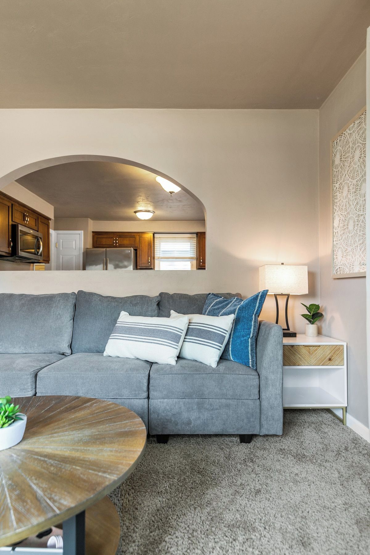 Una sala de estar con un sofá gris, una mesa de centro redonda y una mesita de noche blanca con una lámpara.