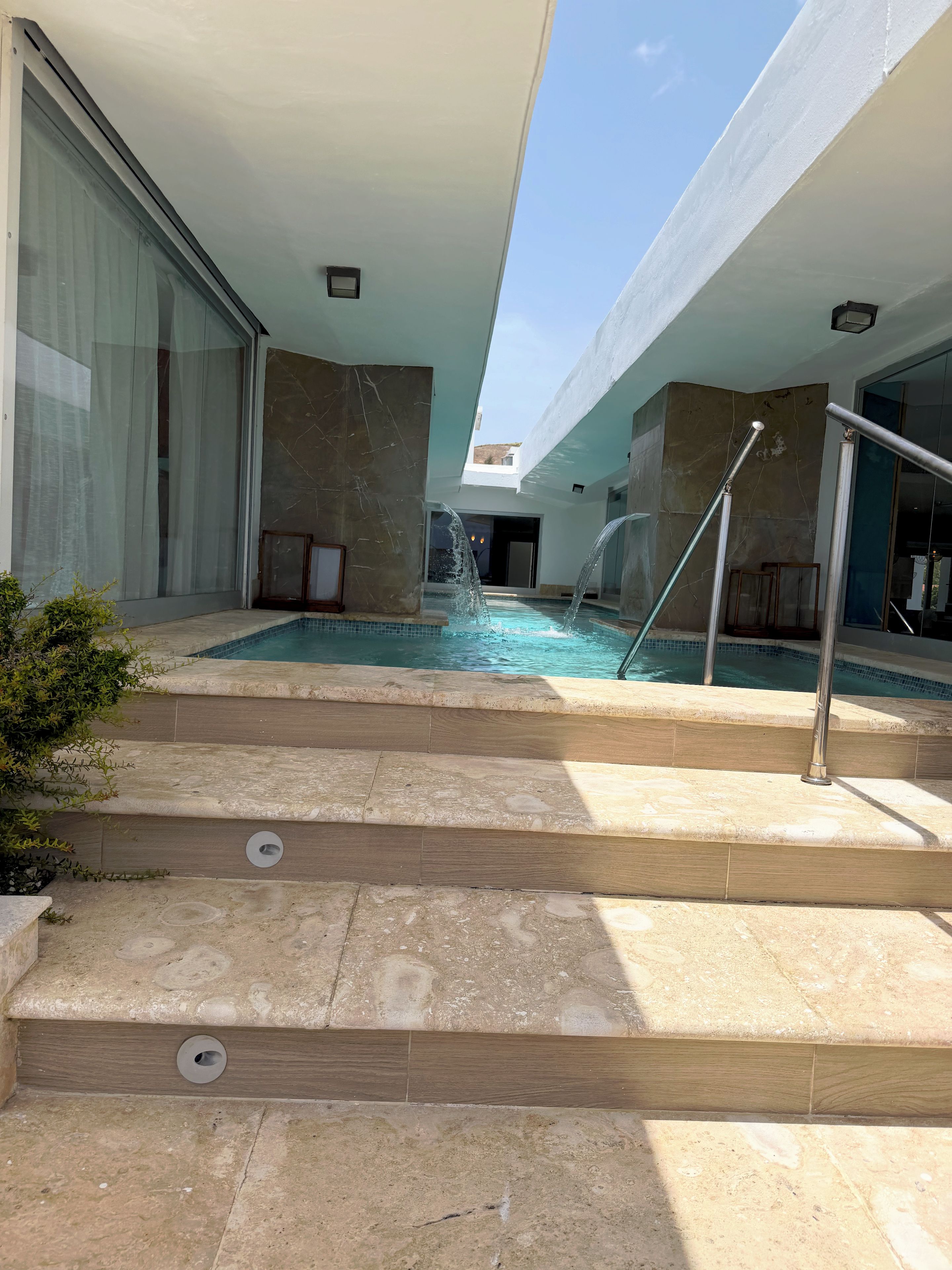 A pool area with steps leading down to the water, a glass door, and a view of the sky.