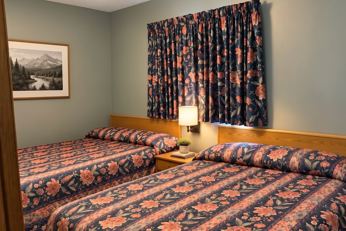 A hotel room with a double bed, a nightstand with a lamp, a framed picture on the wall, and a window with curtains.