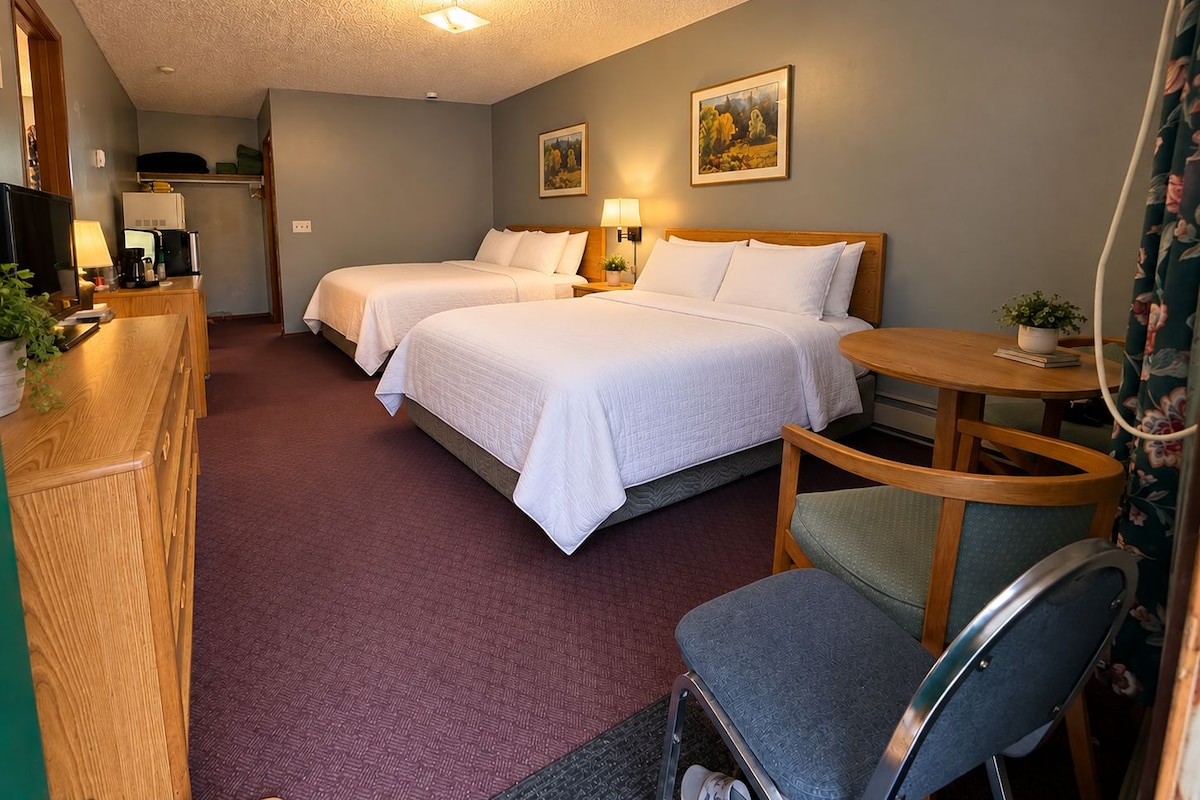A hotel room with two beds, a desk with a lamp, a chair, and a small table with a plant.