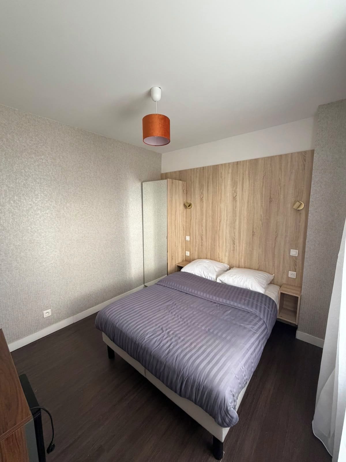 Une chambre à coucher avec un lit, une tête de lit en bois, une table de chevet et un luminaire fixé au mur.