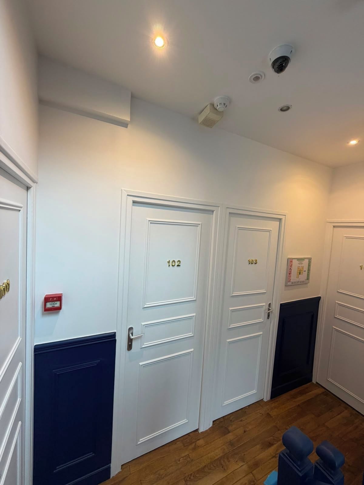 Un couloir avec deux portes blanches, l’une portant le numéro 102 et l’autre le numéro 103, un plancher en bois et un panneau rouge indiquant la sortie de secours.
